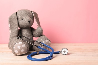 Photo of Pediatrics concept. Toy bunny with stethoscope on wooden table against pink background, space for text