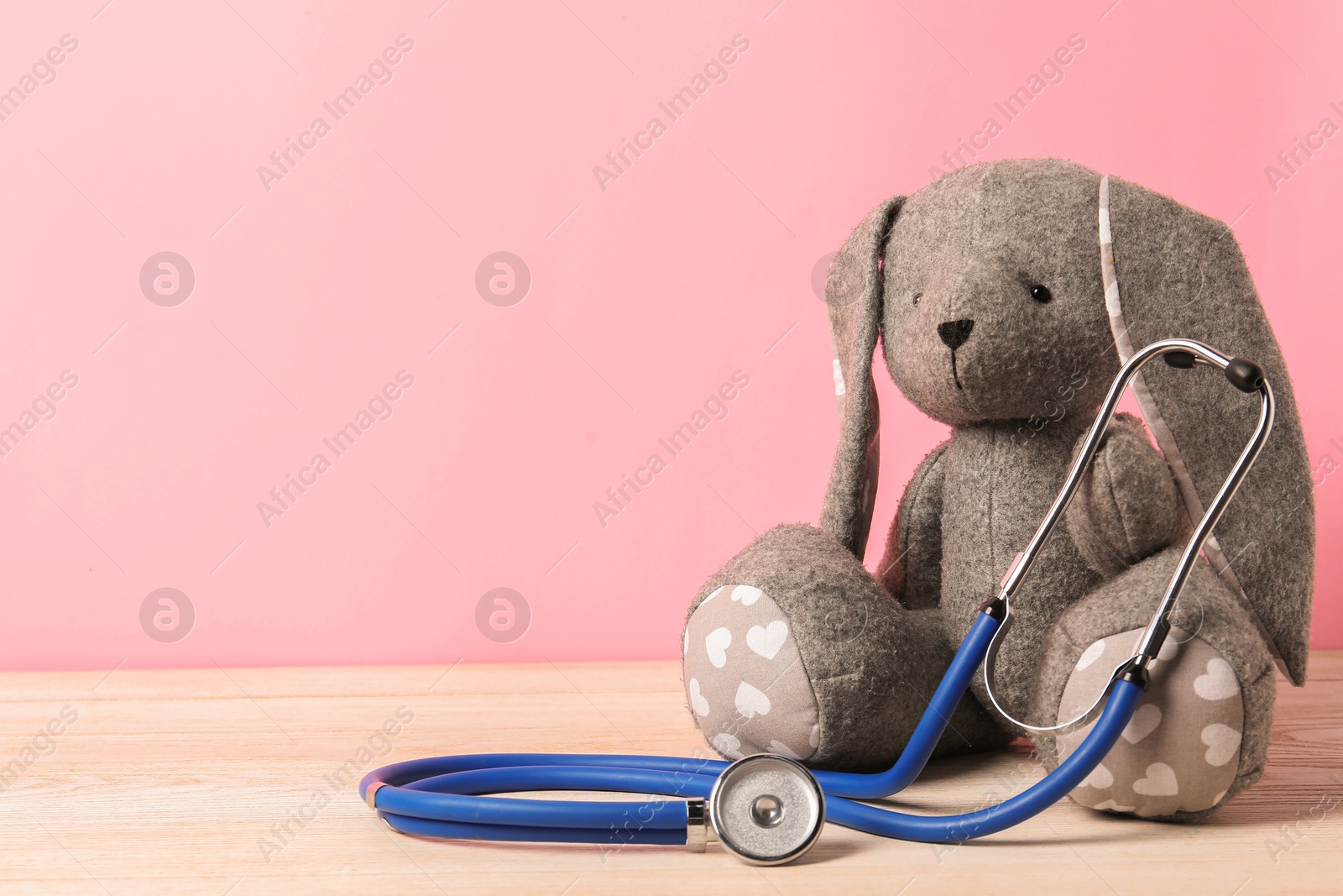 Photo of Pediatrics concept. Toy bunny with stethoscope on wooden table against pink background, space for text