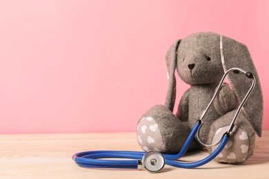 Photo of Pediatrics concept. Toy bunny with stethoscope on wooden table against pink background, space for text