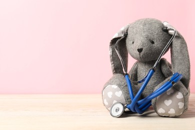 Photo of Pediatrics concept. Toy bunny with stethoscope on wooden table against pink background, space for text