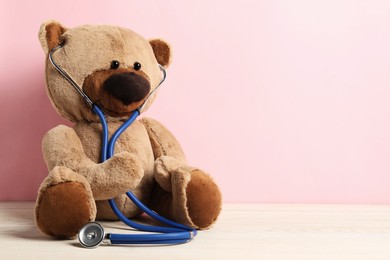 Photo of Pediatrics concept. Teddy bear with stethoscope on wooden table against pink background, space for text