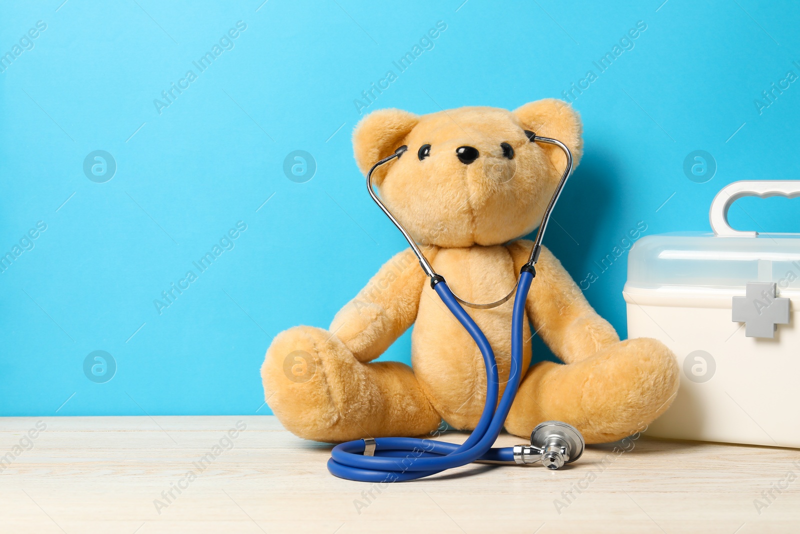 Photo of Pediatrics concept. Teddy bear with stethoscope and first aid kit on wooden table against light blue background, space for text