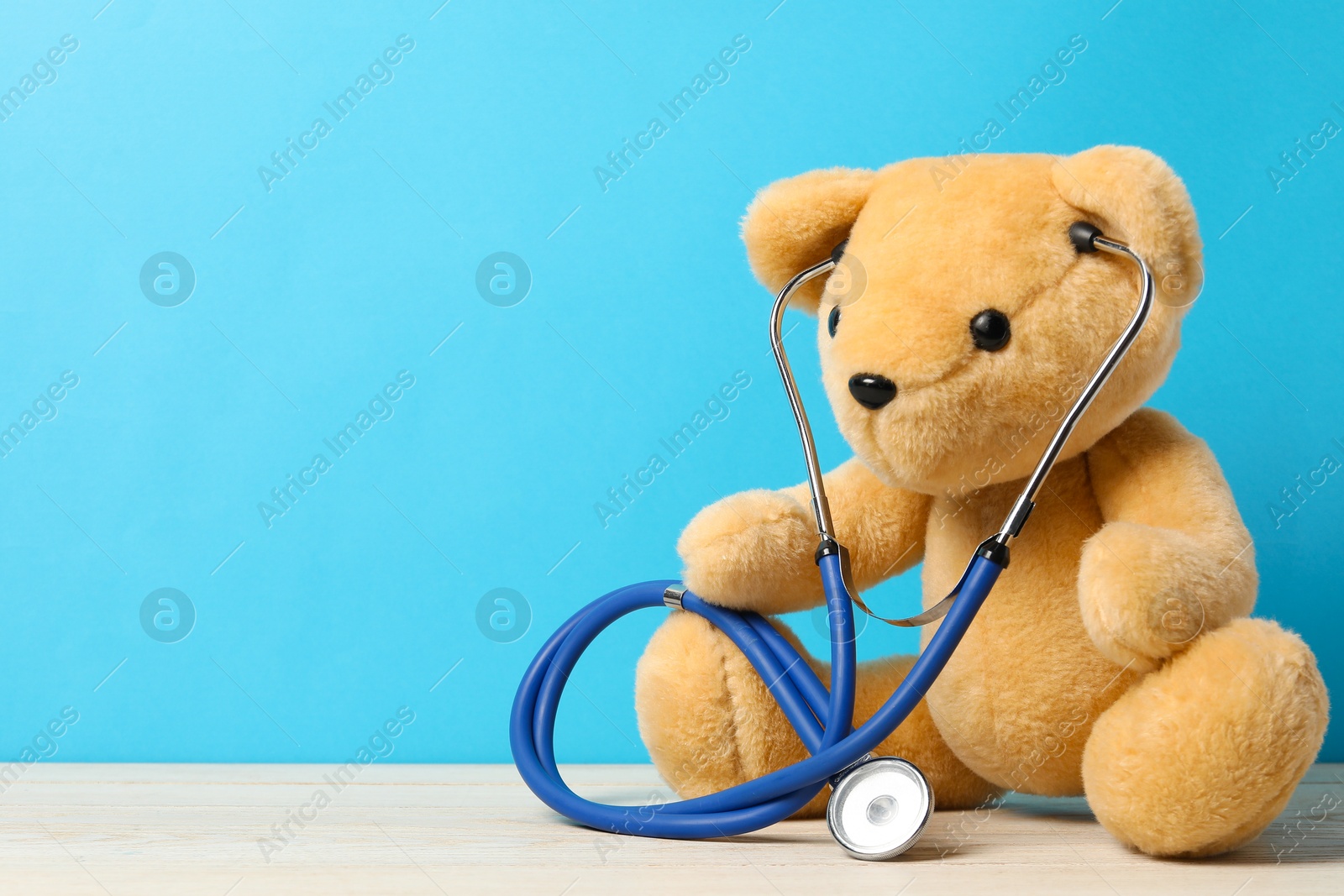 Photo of Pediatrics concept. Teddy bear with stethoscope on wooden table against light blue background, space for text