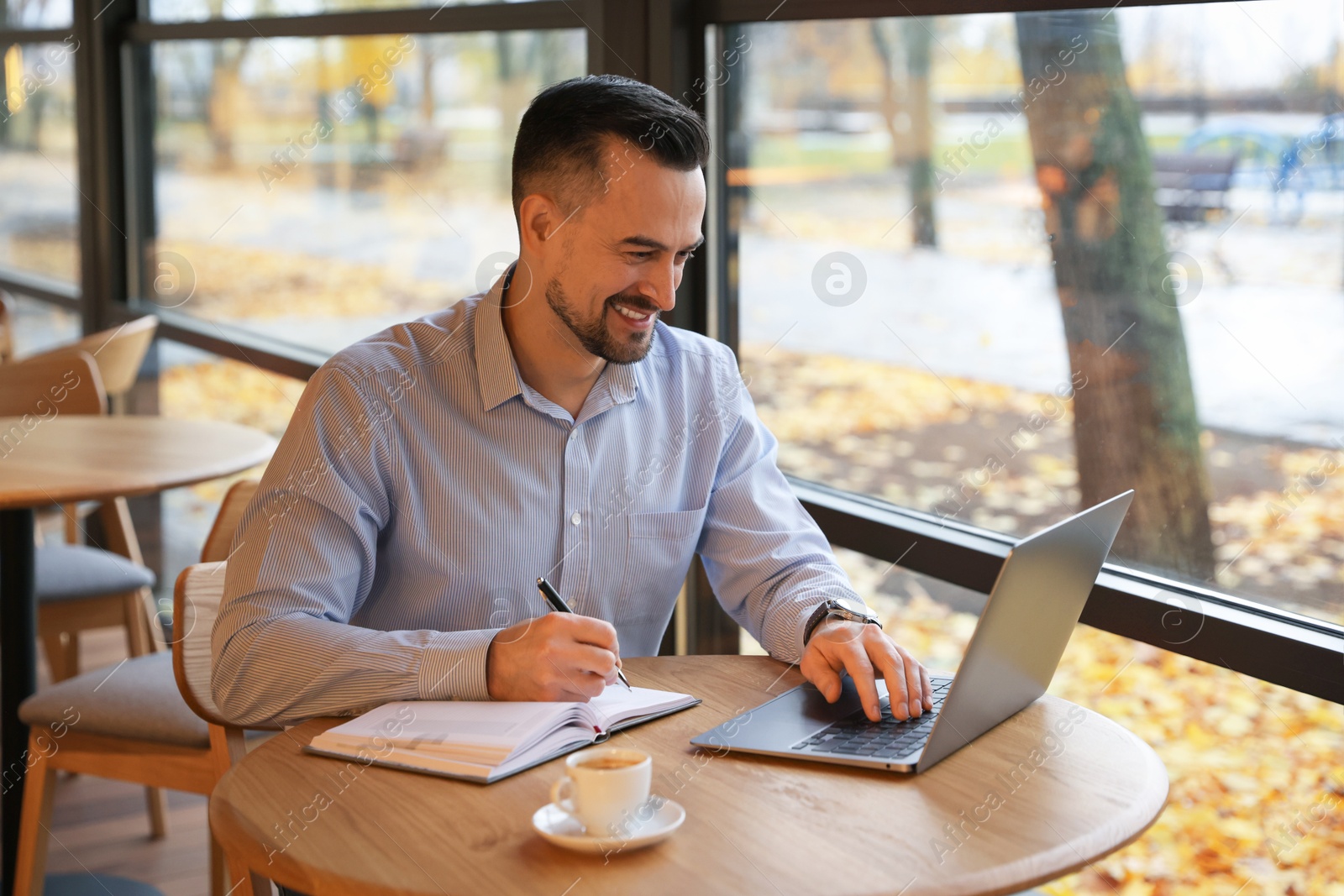 Photo of Man taking notes while working on laptop at table in cafe