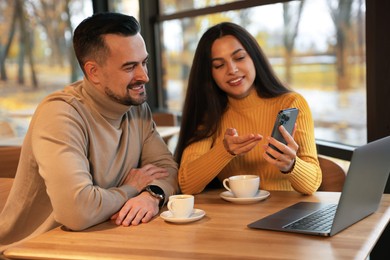 Photo of Woman showing something on smartphone to man at table in cafe