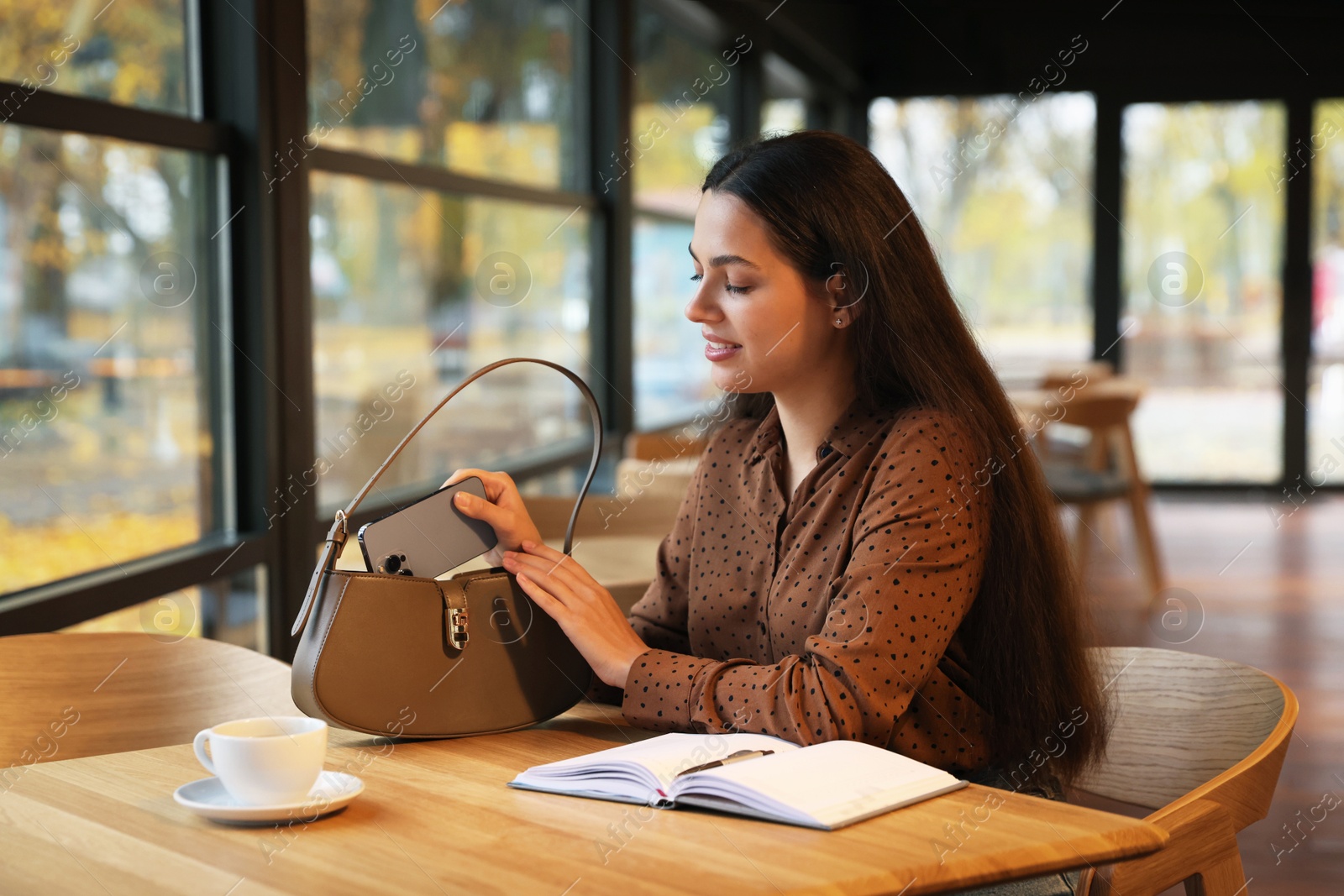 Photo of Woman putting smartphone into bag at table in cafe