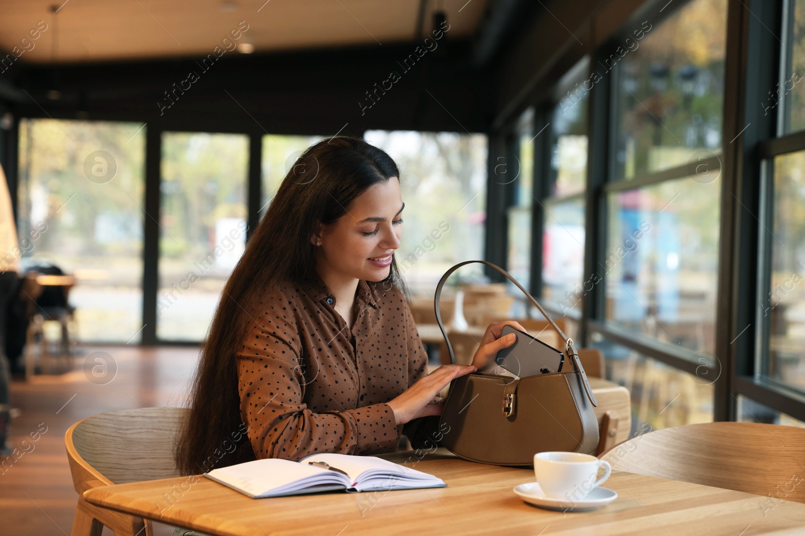 Photo of Woman putting smartphone into bag at table in cafe