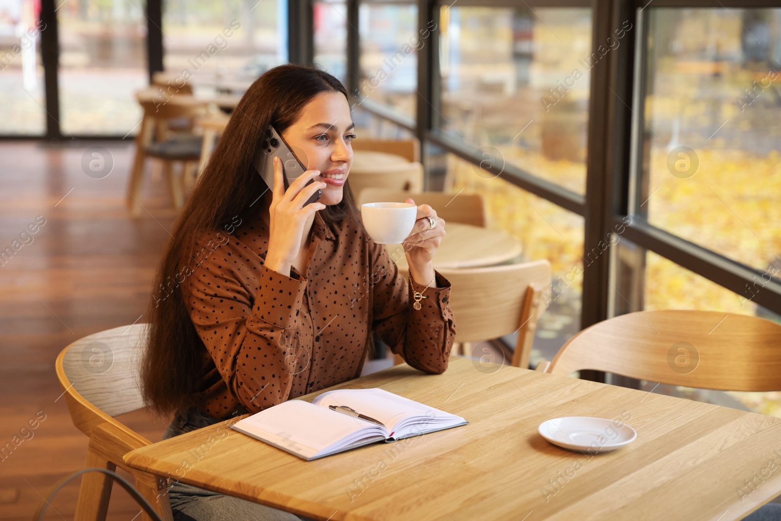 Photo of Woman with cup of coffee talking on smartphone at table in cafe