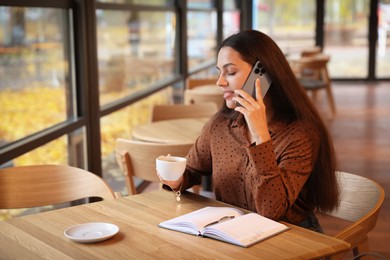 Photo of Woman with cup of coffee talking on smartphone at table in cafe