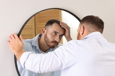 Photo of Baldness problem. Man with receding hairline near mirror at home