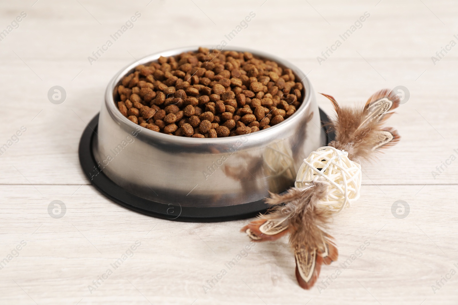 Photo of Dry pet food granules in feeding bowl and cat toy on floor indoors