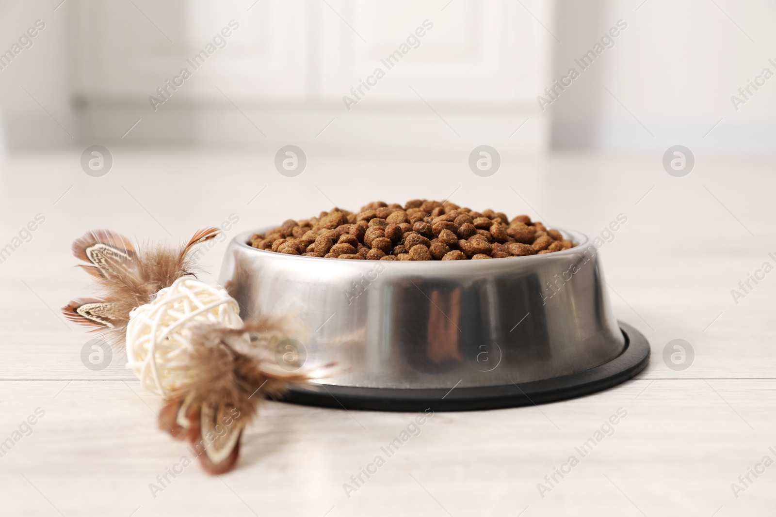 Photo of Dry pet food granules in feeding bowl and cat toy on floor indoors