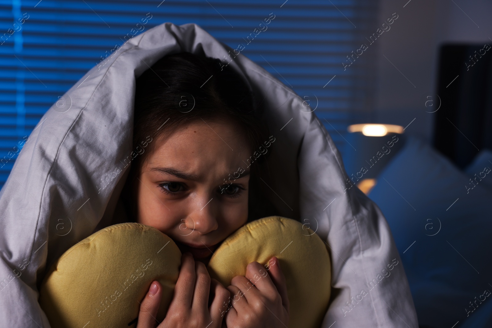 Photo of Afraid girl with pillow under duvet on bed at night