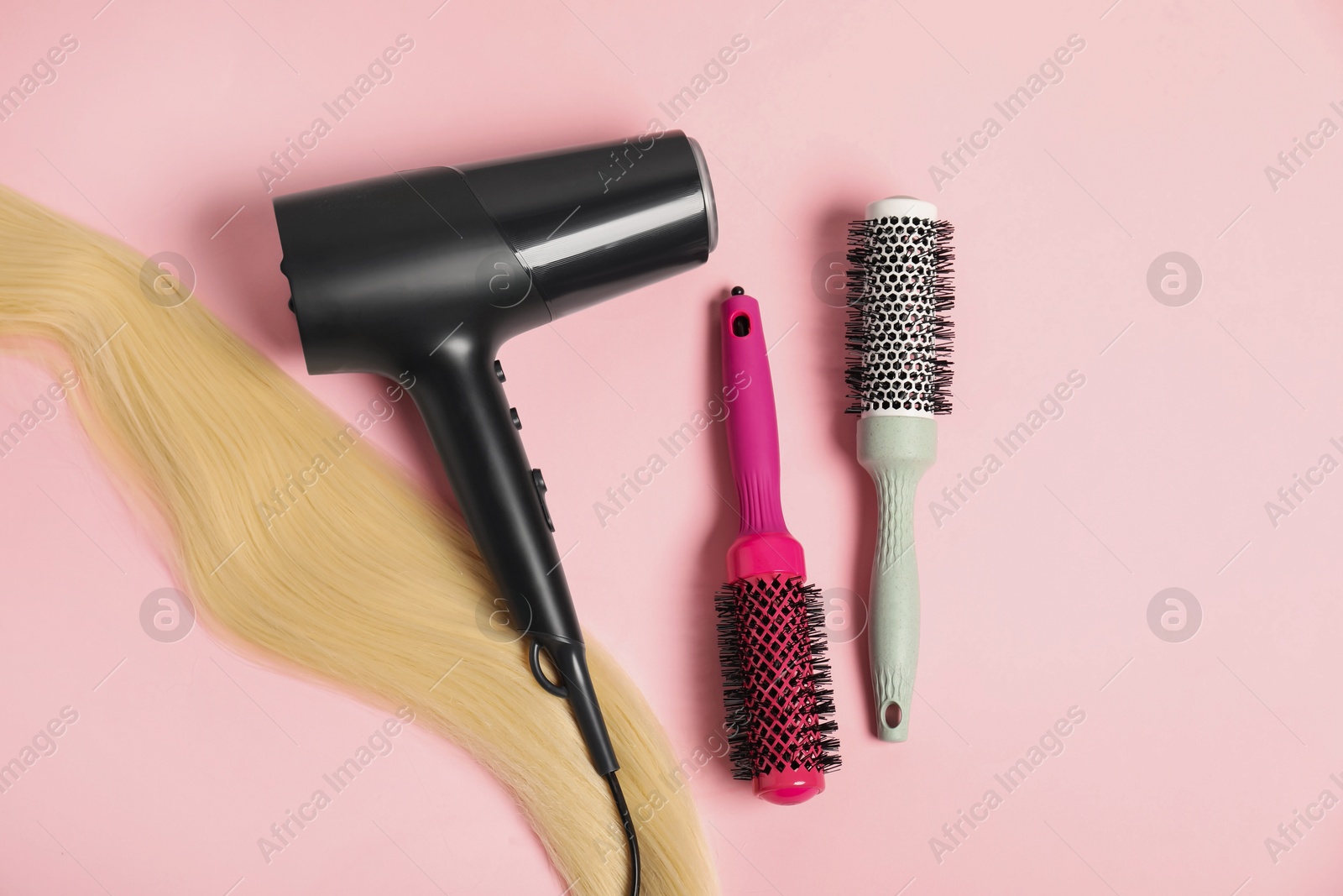 Photo of Stylish round brushes, lock of blonde hair and hairdryer on pink background, flat lay