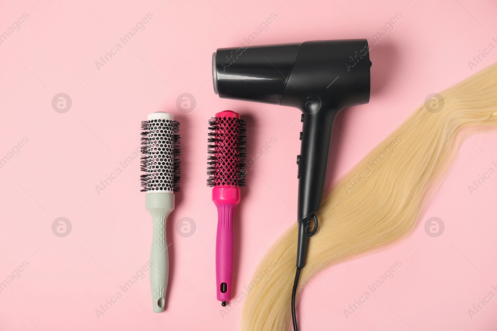 Photo of Stylish round brushes, lock of blonde hair and hairdryer on pink background, flat lay