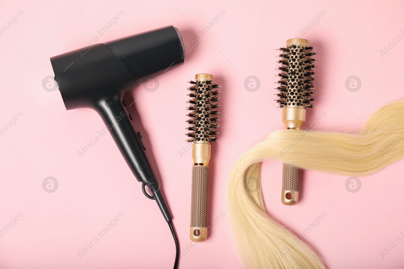 Photo of Stylish round brushes, lock of blonde hair and hairdryer on pink background, flat lay