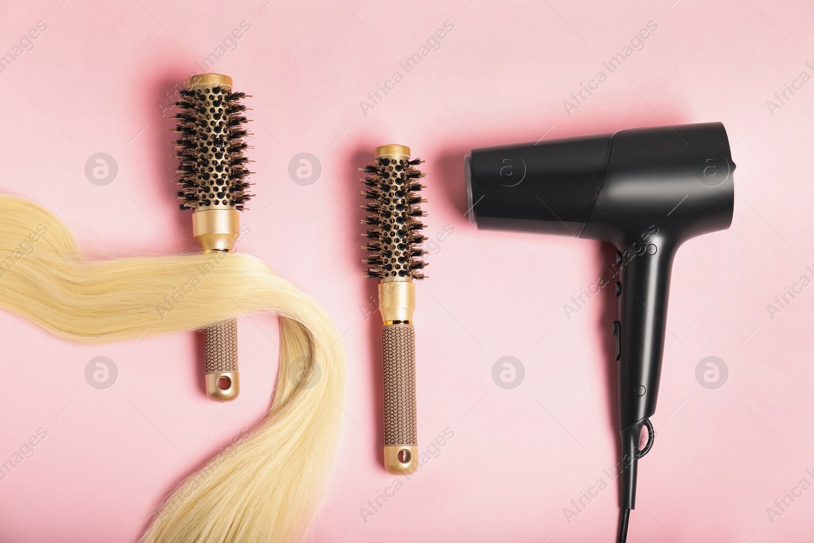 Photo of Stylish round brushes, lock of blonde hair and hairdryer on pink background, flat lay