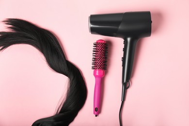 Photo of Stylish round brush, lock of black hair and hairdryer on pink background, flat lay