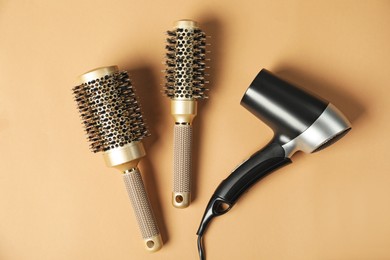 Photo of Stylish round brushes and hairdryer on light brown background, flat lay