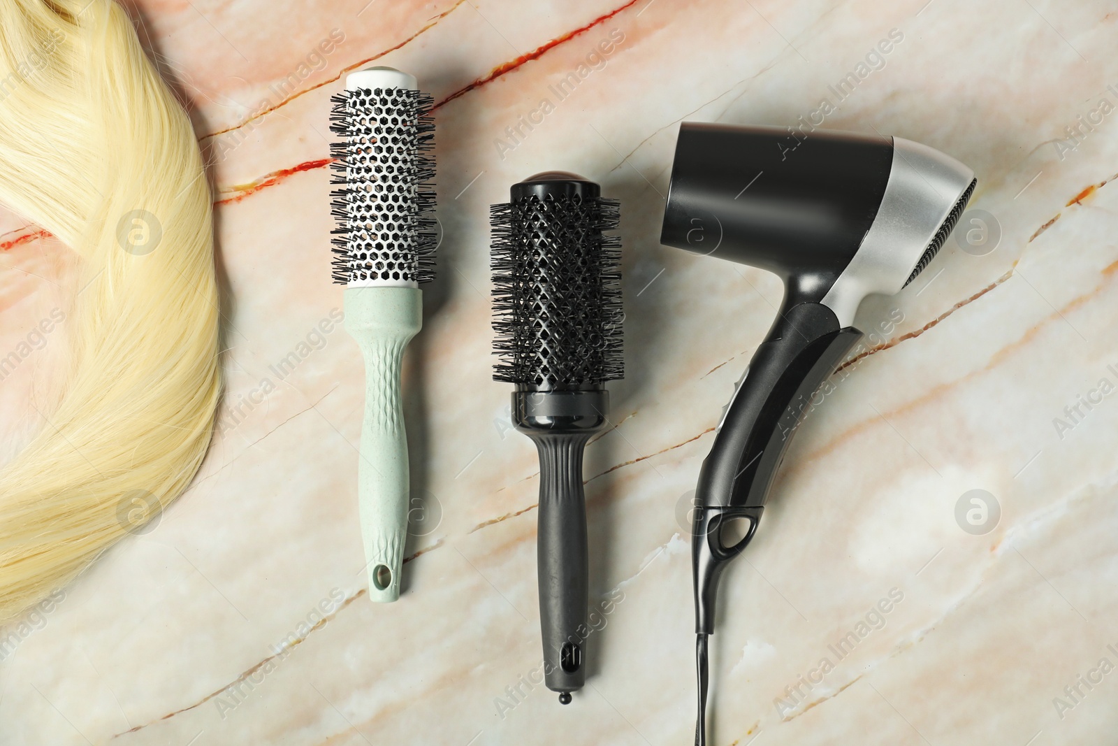 Photo of Stylish round brushes, hairdryer and lock of blonde hair on pink marble table, flat lay