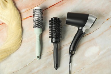 Photo of Stylish round brushes, hairdryer and lock of blonde hair on pink marble table, flat lay