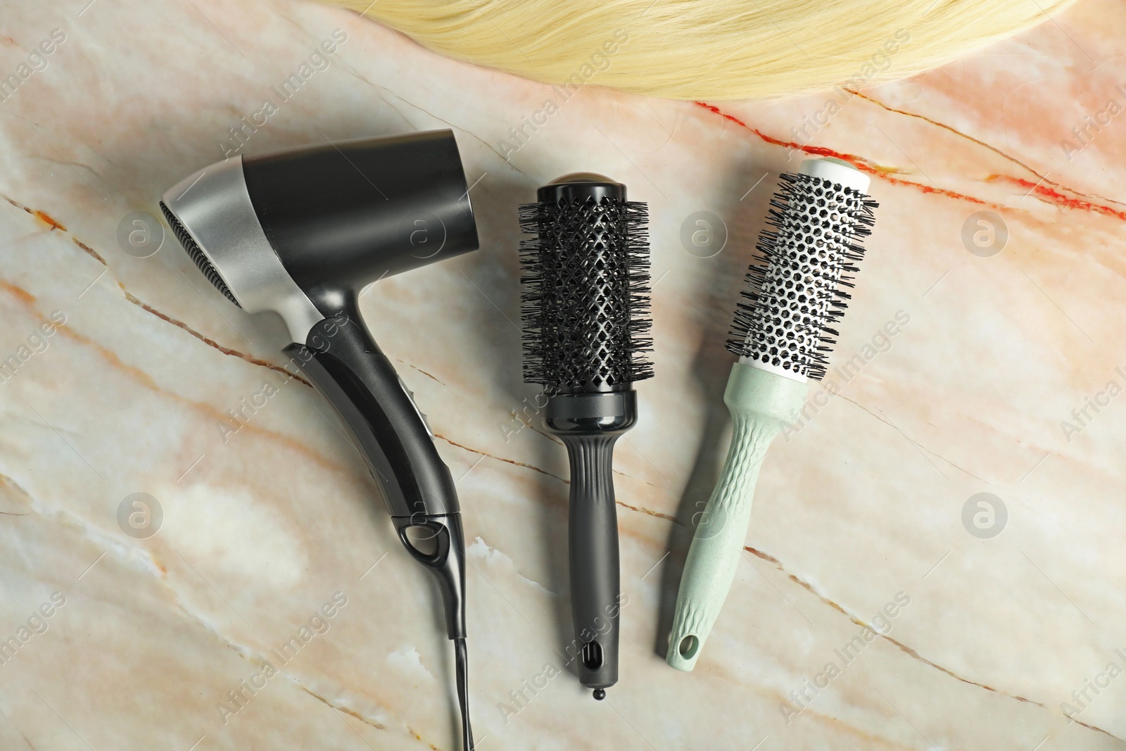 Photo of Stylish round brushes, hairdryer and lock of blonde hair on pink marble table, flat lay