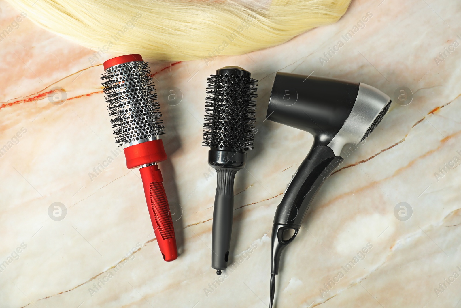 Photo of Stylish round brushes, hairdryer and lock of blonde hair on pink marble table, flat lay