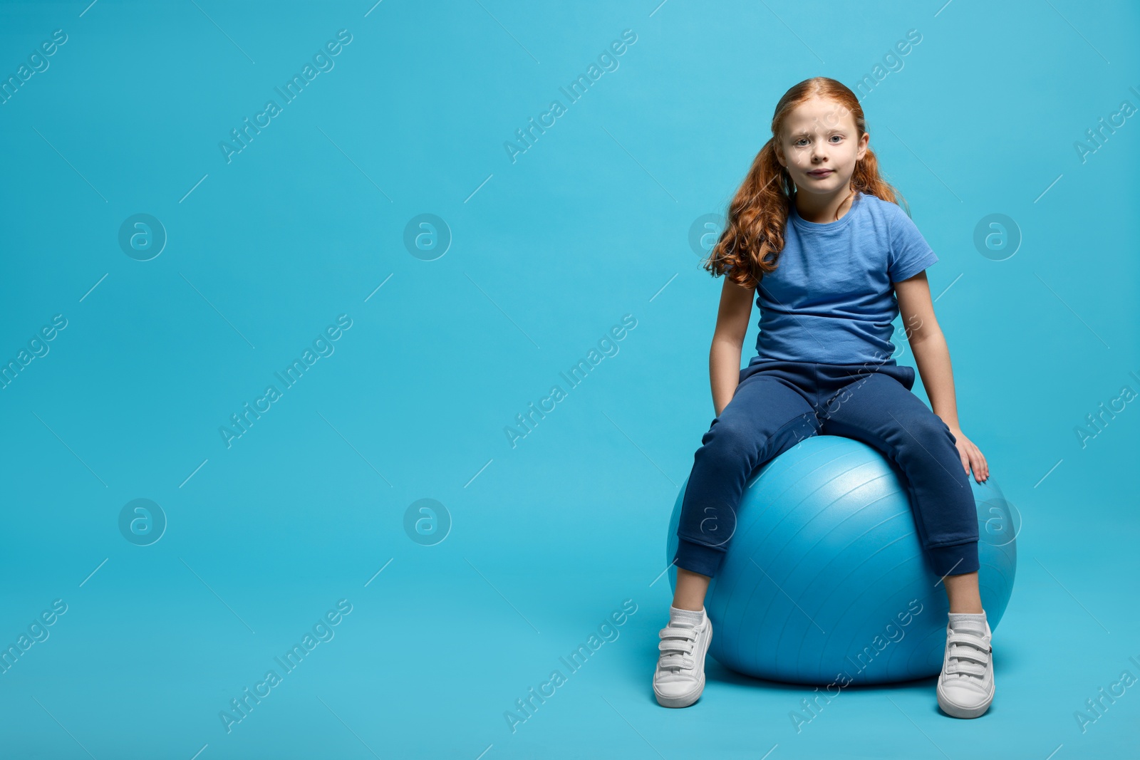 Photo of Cute little girl sitting on fitness ball against light blue background, space for text