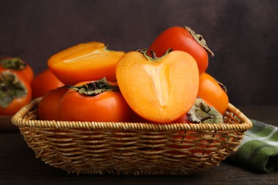 Photo of Whole and cut juicy persimmons in wicker basket on wooden table, closeup