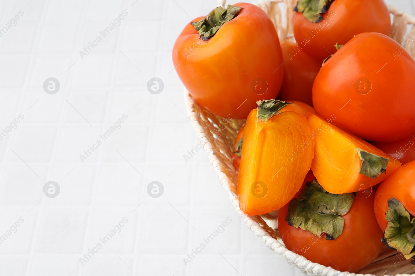 Photo of Ripe persimmons in wicker basket on white tiled table, closeup. Space for text