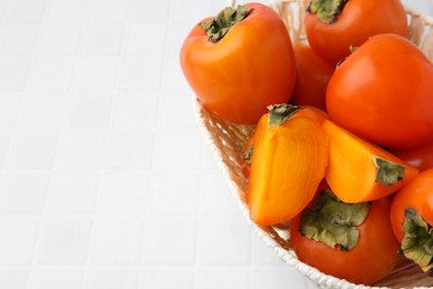 Ripe persimmons in wicker basket on white tiled table, closeup. Space for text