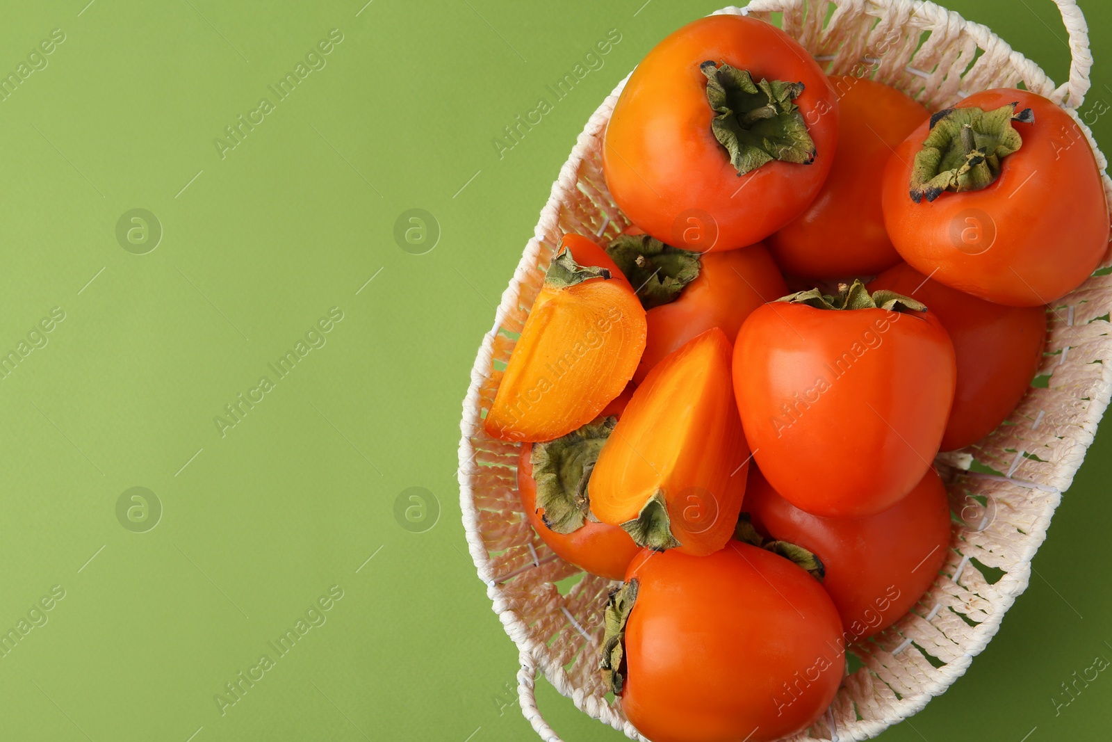 Photo of Ripe persimmons in wicker basket on green background, top view. Space for text