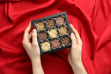 Photo of Woman with box of delicious flower shaped chocolate bonbons on red fabric background, top view