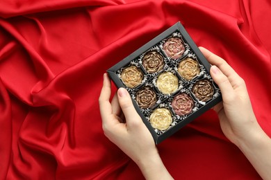 Photo of Woman with box of delicious flower shaped chocolate bonbons on red fabric background, top view. Space for text