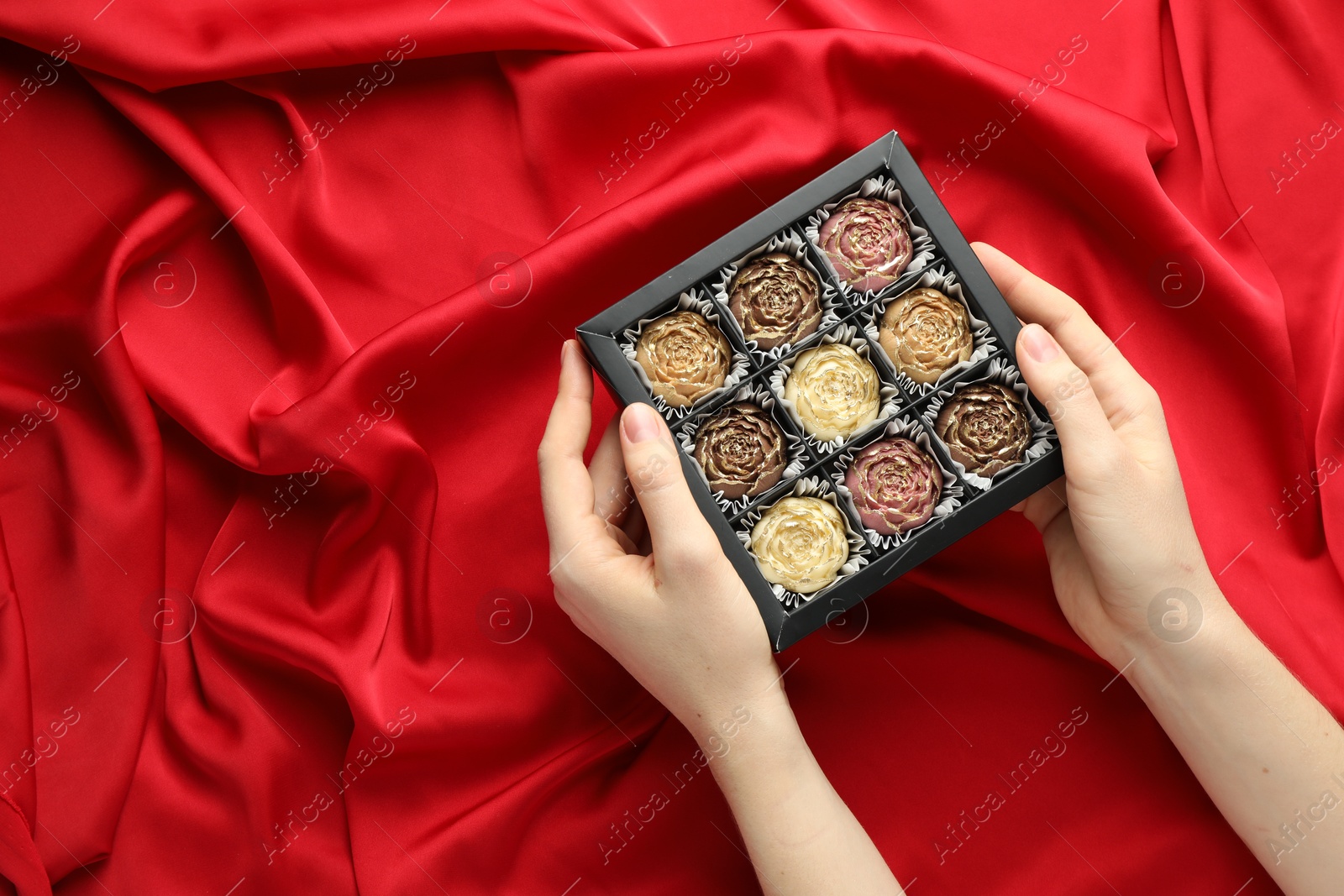 Photo of Woman with box of delicious flower shaped chocolate bonbons on red fabric background, top view. Space for text