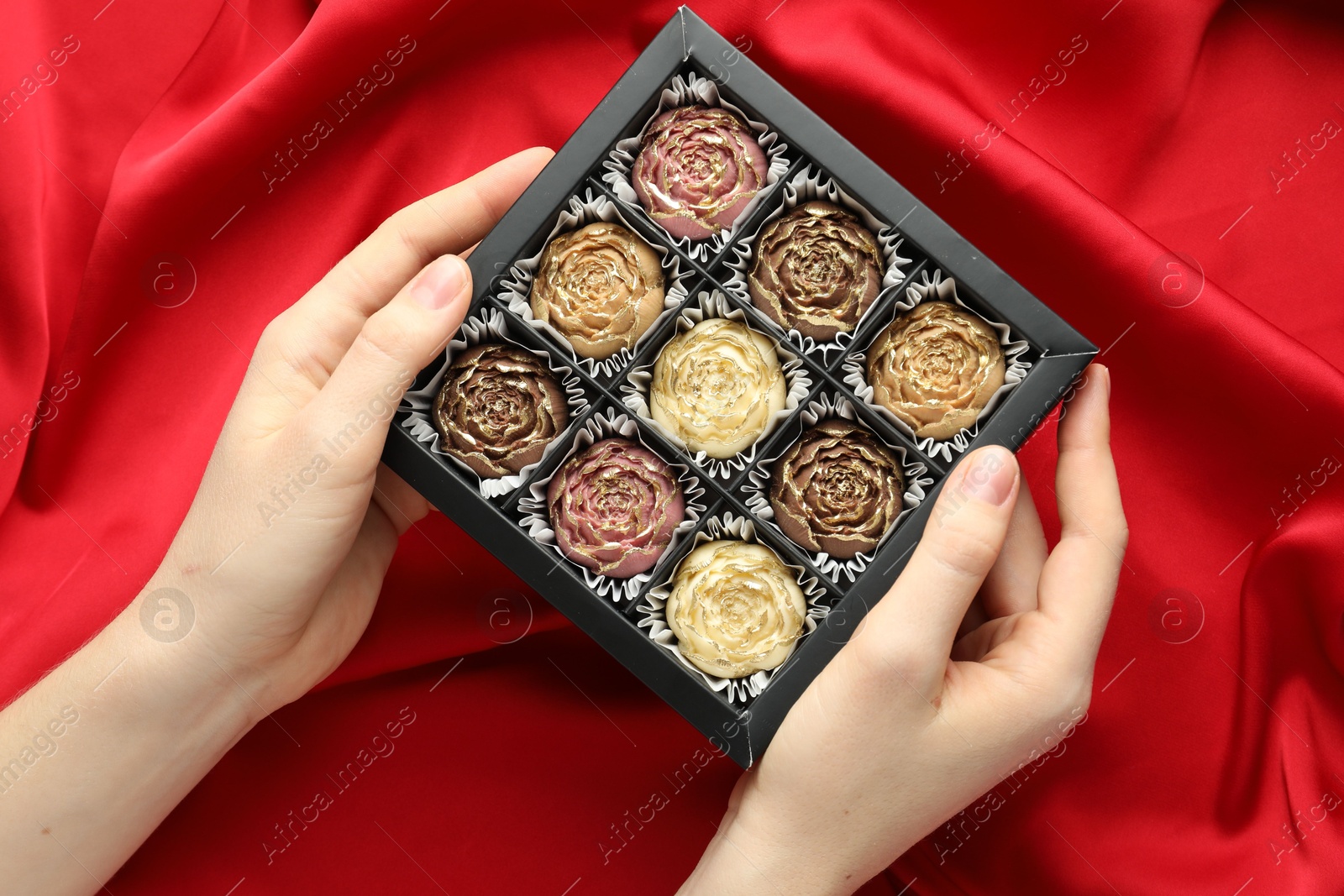 Photo of Woman with box of delicious flower shaped chocolate bonbons on red fabric background, top view