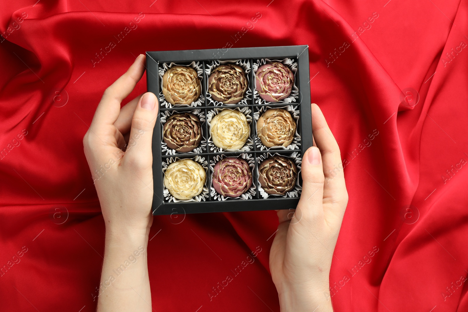 Photo of Woman with box of delicious flower shaped chocolate bonbons on red fabric background, top view