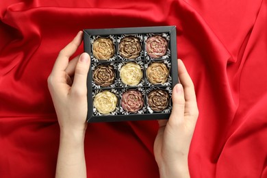 Photo of Woman with box of delicious flower shaped chocolate bonbons on red fabric background, top view