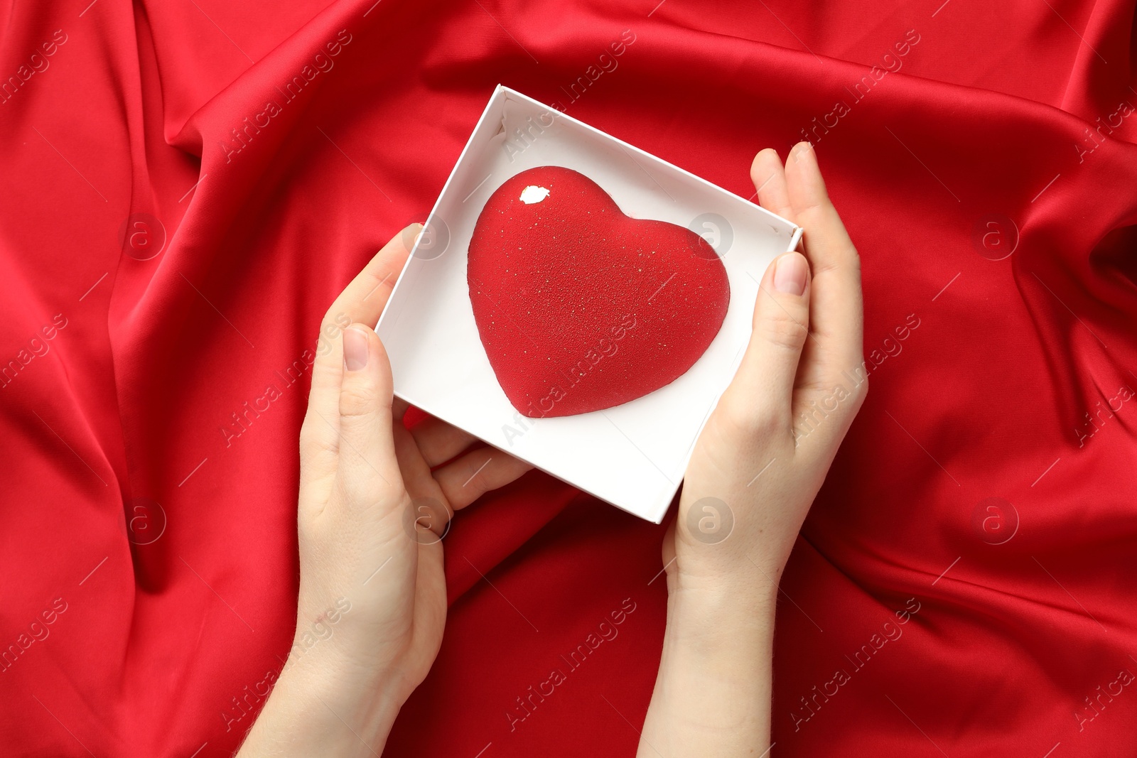 Photo of Woman with heart shaped chocolate in box on red fabric background, top view