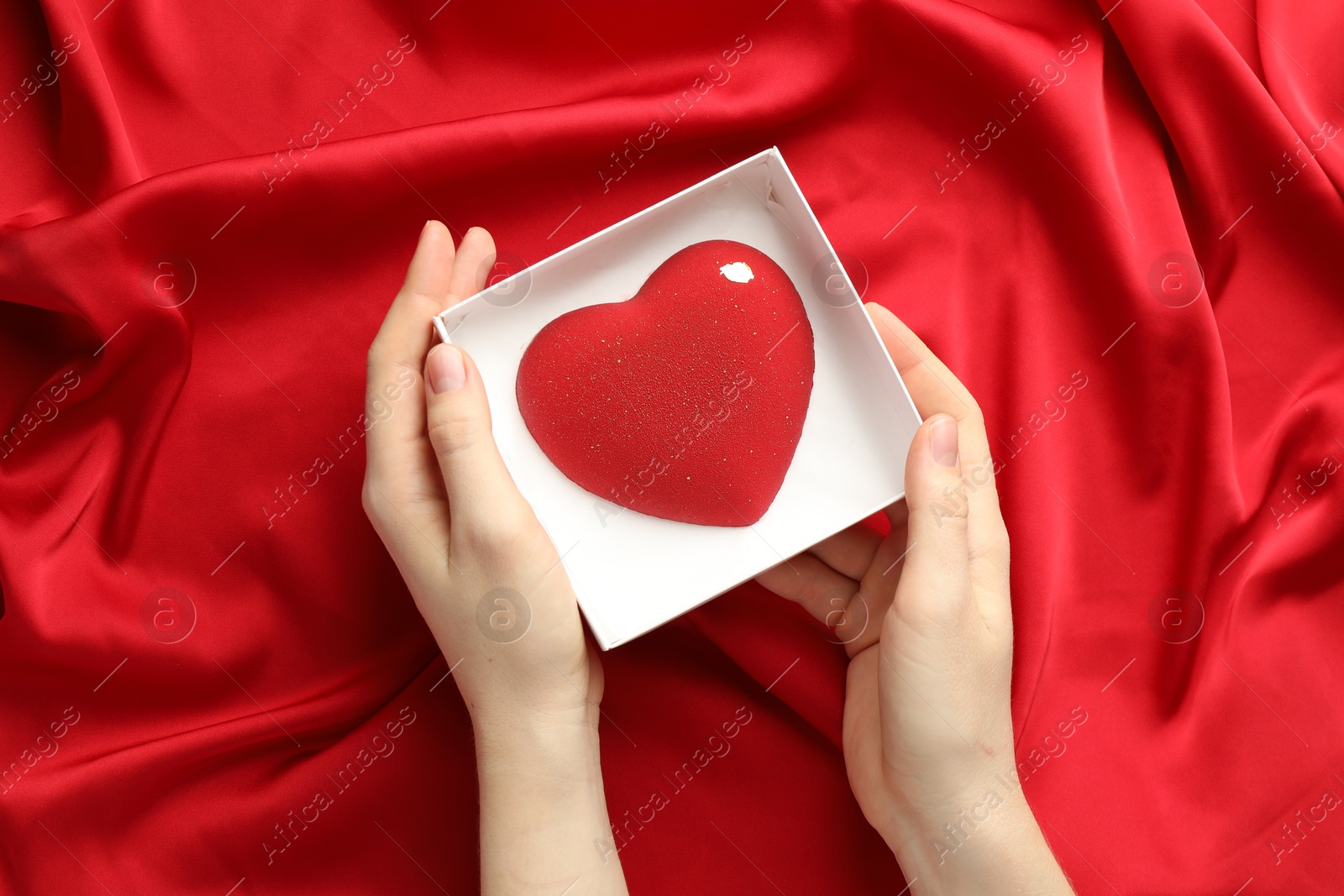 Photo of Woman with heart shaped chocolate in box on red fabric background, top view