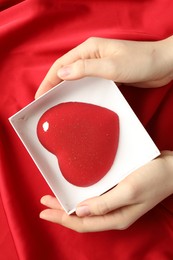 Photo of Woman with heart shaped chocolate in box on red fabric background, top view