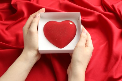 Photo of Woman with heart shaped chocolate in box on red fabric background, closeup