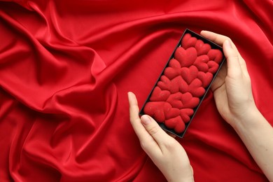 Photo of Woman holding chocolate bar decorated with hearts on red fabric background, top view. Space for text