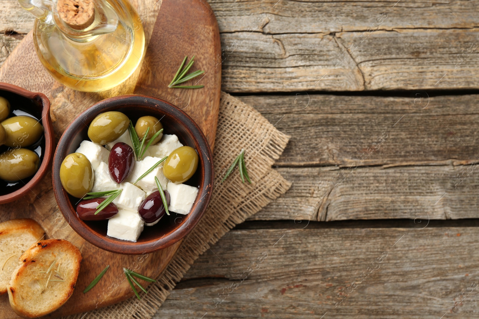 Photo of Marinated olives with feta cheese, bread pieces, oil and rosemary on wooden table, top view. Space for text