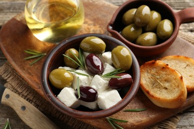 Photo of Marinated olives with feta cheese, bread pieces, oil and rosemary on wooden table, closeup