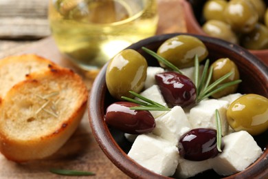 Photo of Marinated olives with feta cheese, bread pieces, oil and rosemary on wooden table, closeup