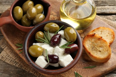 Photo of Marinated olives with feta cheese, bread pieces, oil and rosemary on wooden table, closeup