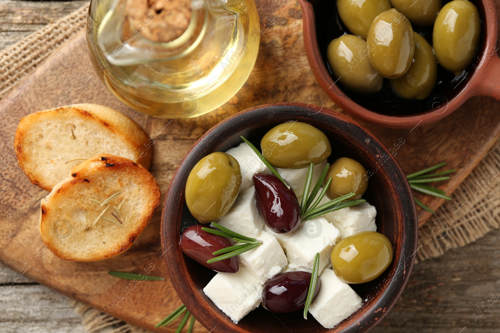 Photo of Marinated olives with feta cheese, bread pieces, oil and rosemary on wooden table, top view