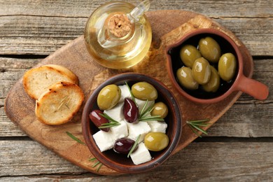 Photo of Marinated olives with feta cheese, bread pieces, oil and rosemary on wooden table, top view