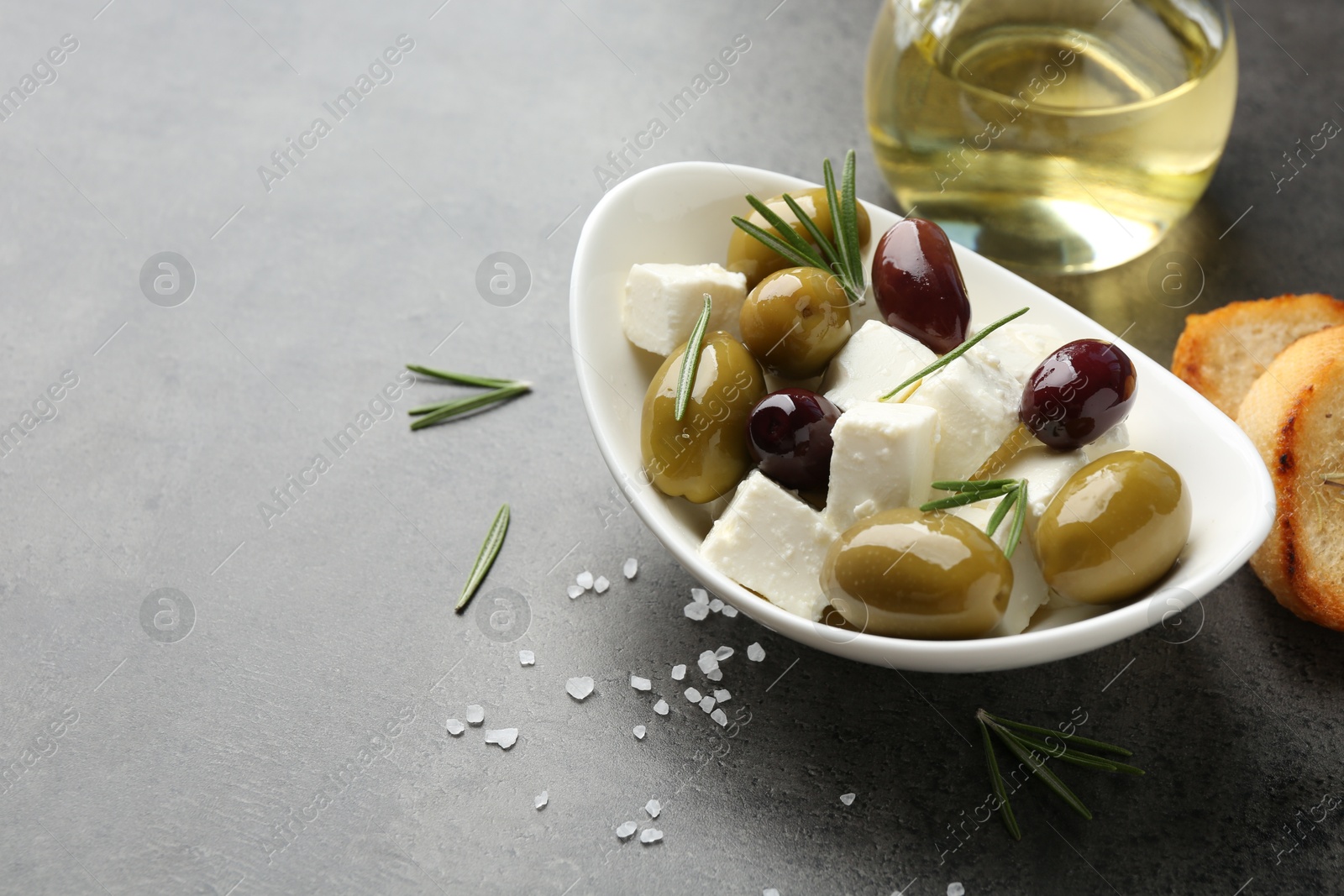 Photo of Marinated olives with feta cheese, bread pieces, oil, salt and rosemary on grey table, closeup. Space for text